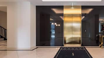 Un ascenseur moderne aux finitions métalliques, un tapis à motifs noirs et dorés et un escalier situé à gauche.