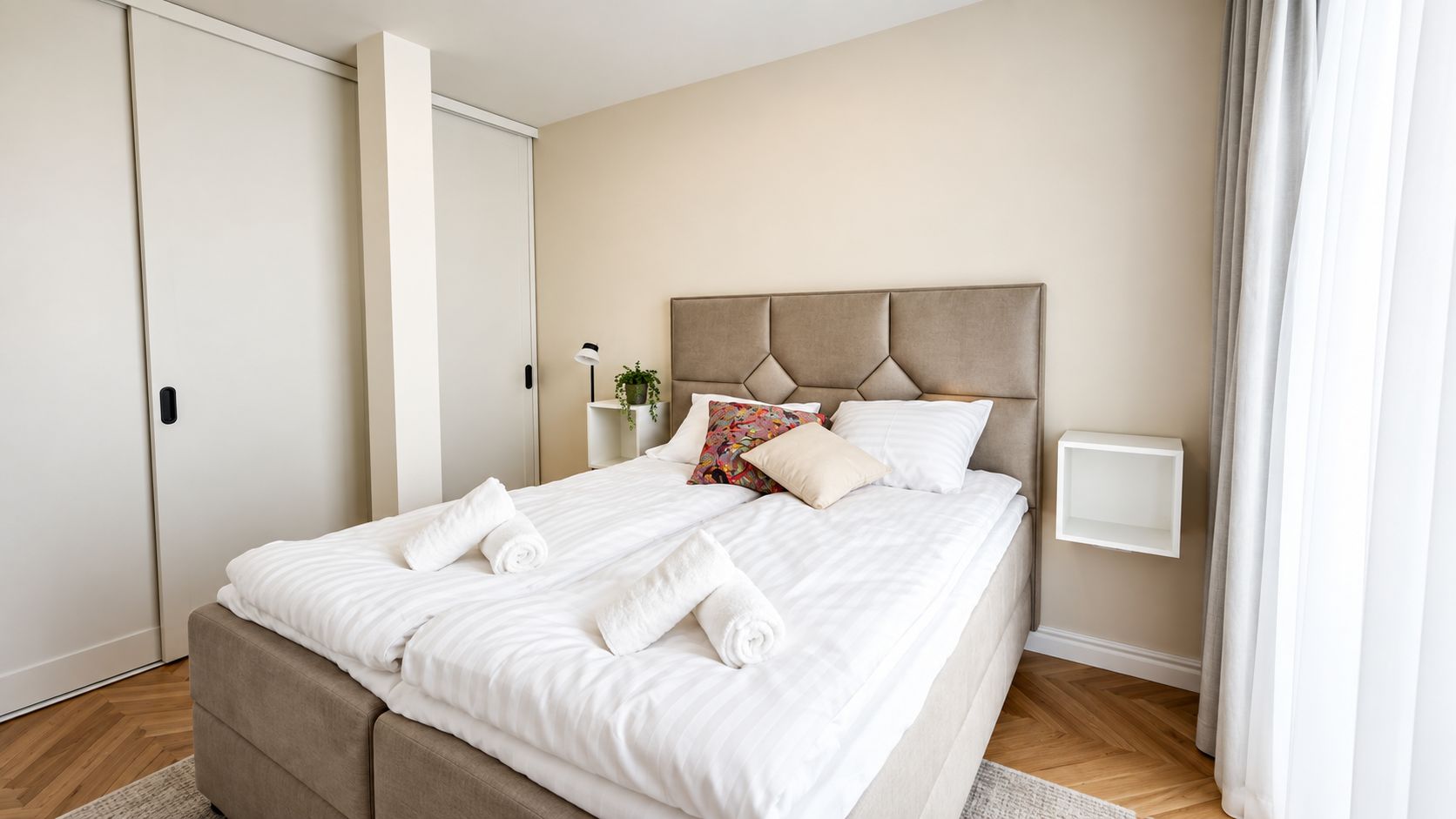 Un lit bien fait, avec du linge de lit blanc et une tête de lit, une table de chevet avec une lampe et une fenêtre avec des rideaux.