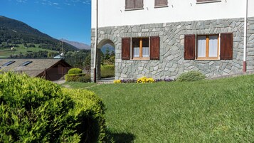 Un edificio in pietra con tetto spiovente, un piccolo giardino fiorito e montagne sullo sfondo.