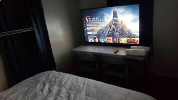 A hotel room with a bed, a desk with a computer, and a TV displaying a movie poster.