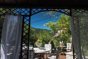 A covered outdoor seating area with a table and chairs, surrounded by greenery and a view of hills. - Anerada Inn Suites Pool & Spa | Lilac Suite (Megalo chorio)