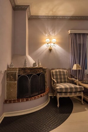 A room with a fireplace, a plaid armchair, a table with a lamp, and a wall-mounted light fixture. - Anerada Inn Suites Pool & Spa | Lilac Suite (Megalo chorio)