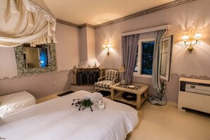 A hotel room with a large bed, a fireplace, a chair, a small table with a vase of flowers, and a nightstand. - Anerada Inn Suites Pool & Spa | Lilac Suite (Megalo chorio)