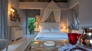 A bedroom with a canopy bed, bedside lamps, a view of greenery outside, and a glass table with decorative items. - Anerada Inn Suites Pool & Spa | Blue Suite (Megalo chorio)