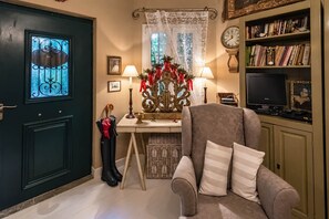 A cozy living room with a dark door, a chair with striped cushions, a table with a decorative centerpiece, and a bookshelf with books and decorative items. - Anerada Inn Suites Pool & Spa | Garden House (Megalo chorio)