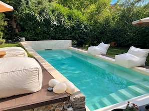 Ein rechteckiger Swimmingpool mit Steinkante, umgeben von einer Holzterrasse und weißen Gartenmöbeln.