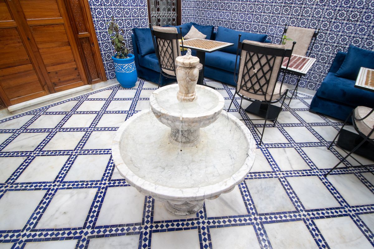 Une cour avec une fontaine, des carreaux bleus à motifs, une porte en bois et des canapés bleus ornés d’un motif d’échiquier.
