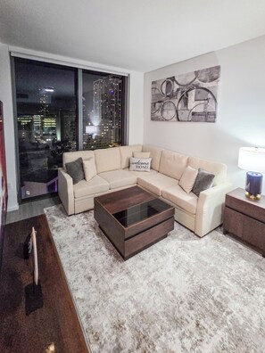 A modern living room with a sectional sofa, a glass coffee table, and a city view through large windows. - Luxury Apartment with shared pool (Miami)