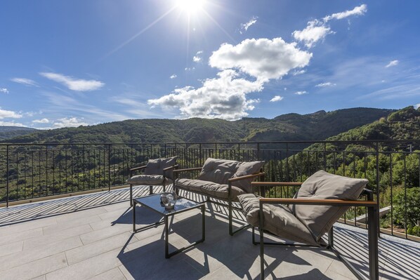 A terrace with a sofa and chairs, a glass table, and a view of a mountainous landscape.