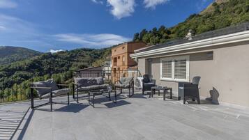 A terrace with outdoor furniture, a view of a hillside, and a building with a sloped roof.