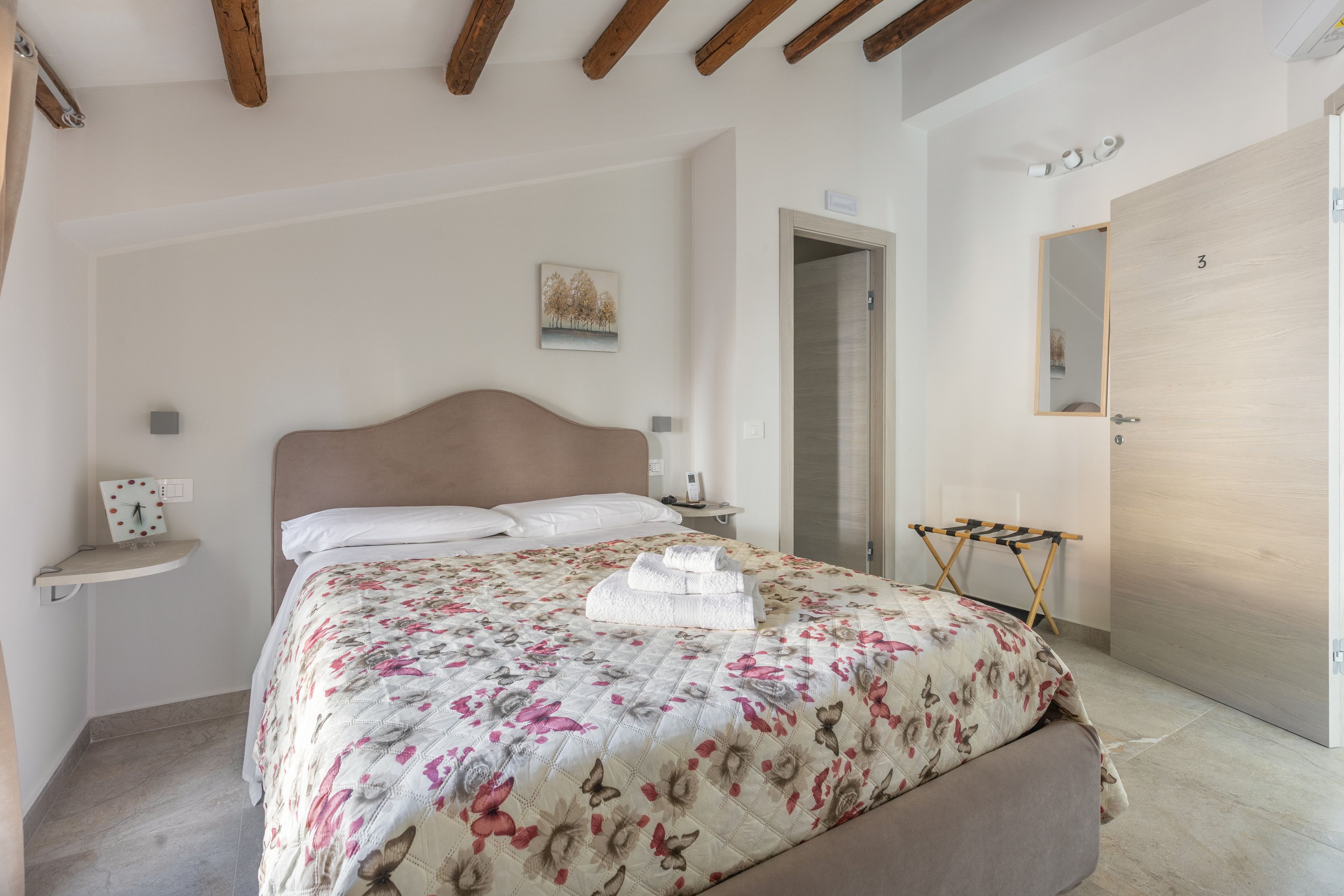 A bedroom with a bed, a nightstand, a bedside table, a wall-mounted clock, a door, and a mirror.