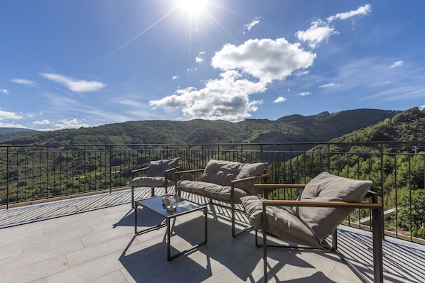 A terrace with a sofa and chairs, a glass table, and a view of a mountainous landscape.