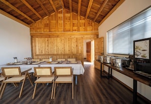 Un comedor con techo alto de madera, una larga mesa dispuesta para una comida, y una zona de cocina con ollas y sartenes.