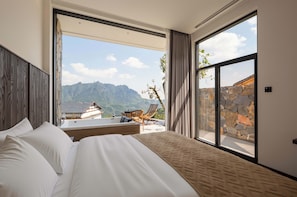 Habitación de hotel con una cama grande, vistas a las montañas y un balcón con mesa y sillas.