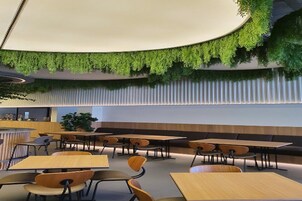 Un interior moderno con mesas y sillas redondas, techo curvo con plantas y una zona de estar con bancos.