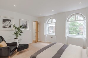 Une chambre d’hôtel avec un lit, une chaise, une plante en pot et deux fenêtres donnant sur un bâtiment en briques et sur de la verdure.