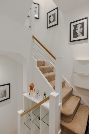 Un escalier avec une rampe en bois, des marches recouvertes d’un tapis beige et des murs blancs. Des tableaux encadrés ornent les murs, et une petite étagère expose quelques objets décoratifs.