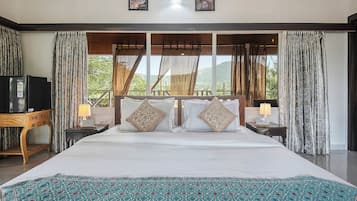 Un dormitorio amplio con una cama grande, dos mesitas de noche, un televisor y vistas a zonas verdes por las ventanas.