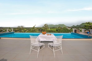 A dining area with a table and chairs facing a pool, with a view of hills in the background.