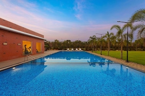 A swimming pool with a clear blue surface, surrounded by a brick building with a covered area, and palm trees in the background. - Fortune Park Airport Road Hubballi (Hubli)