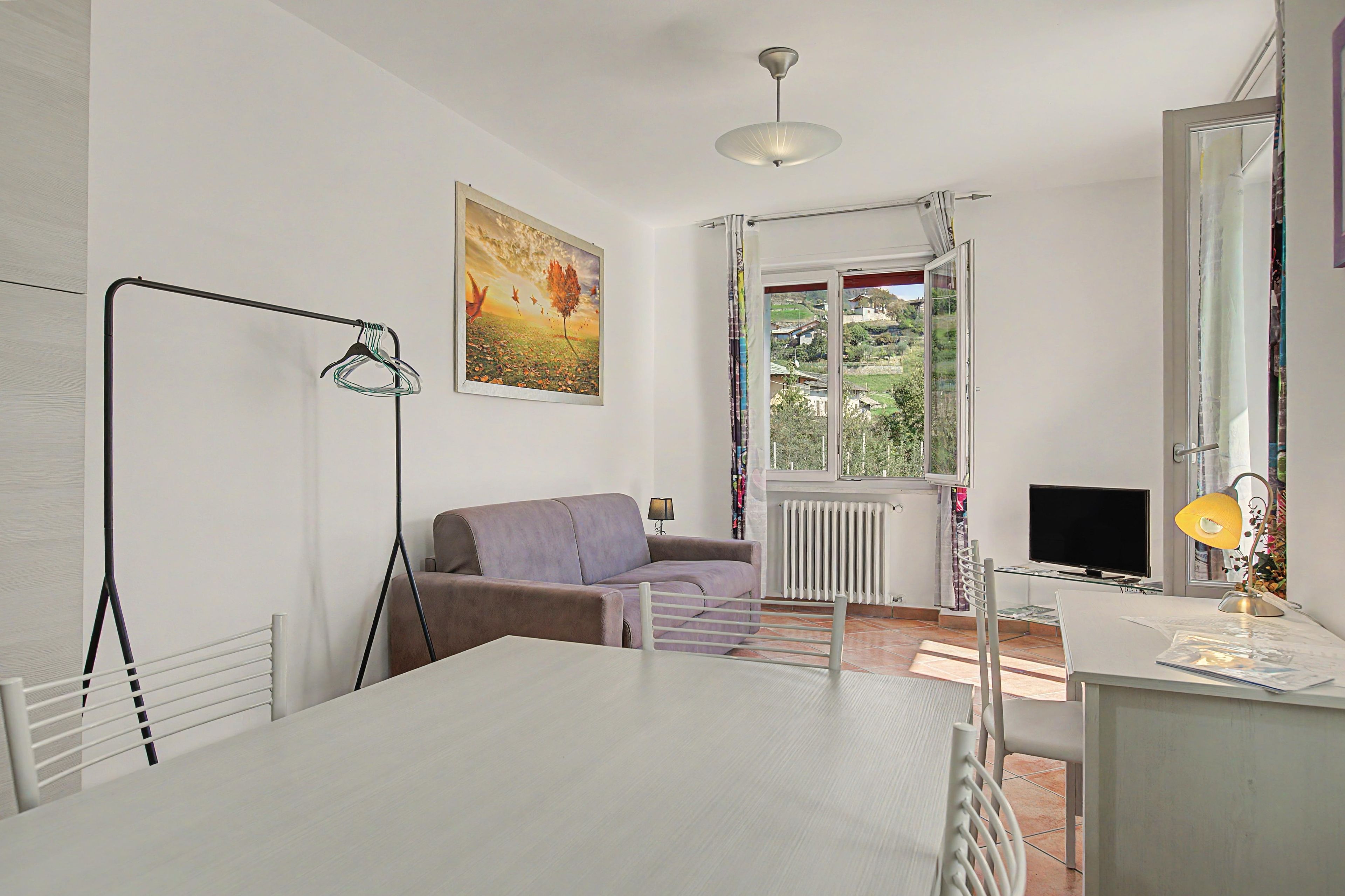 Ein Zimmer mit einem weißen Tisch, einem violetten Sofa, einem Heizkörper, einem Fenster mit Blick auf Grünflächen und einer Deckenleuchte.