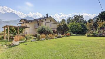 Une maison avec un jardin, une pergola et des montagnes en arrière-plan.