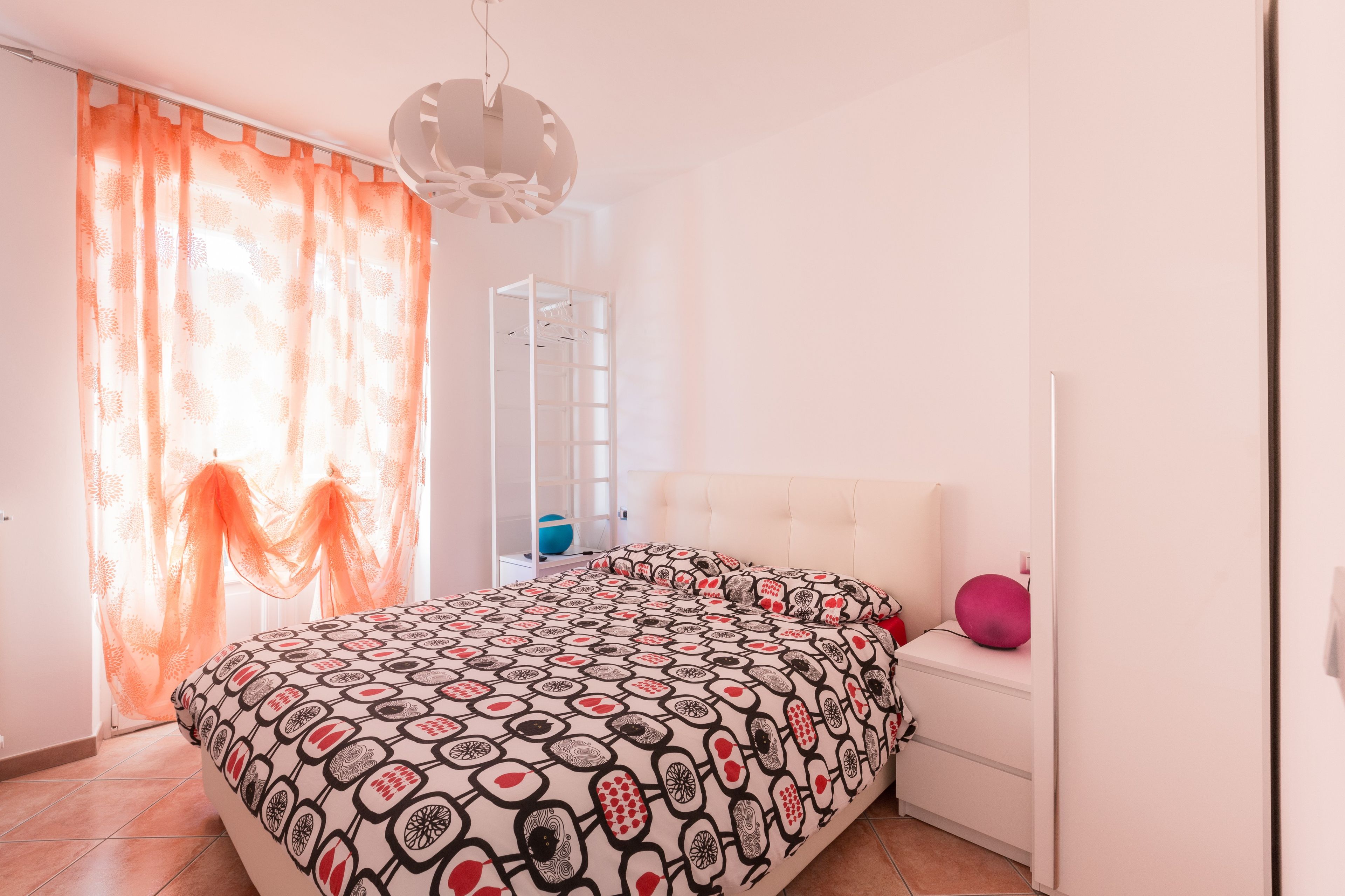 A bedroom with a bed, a bedside table, a hanging light fixture, and a window with curtains.
