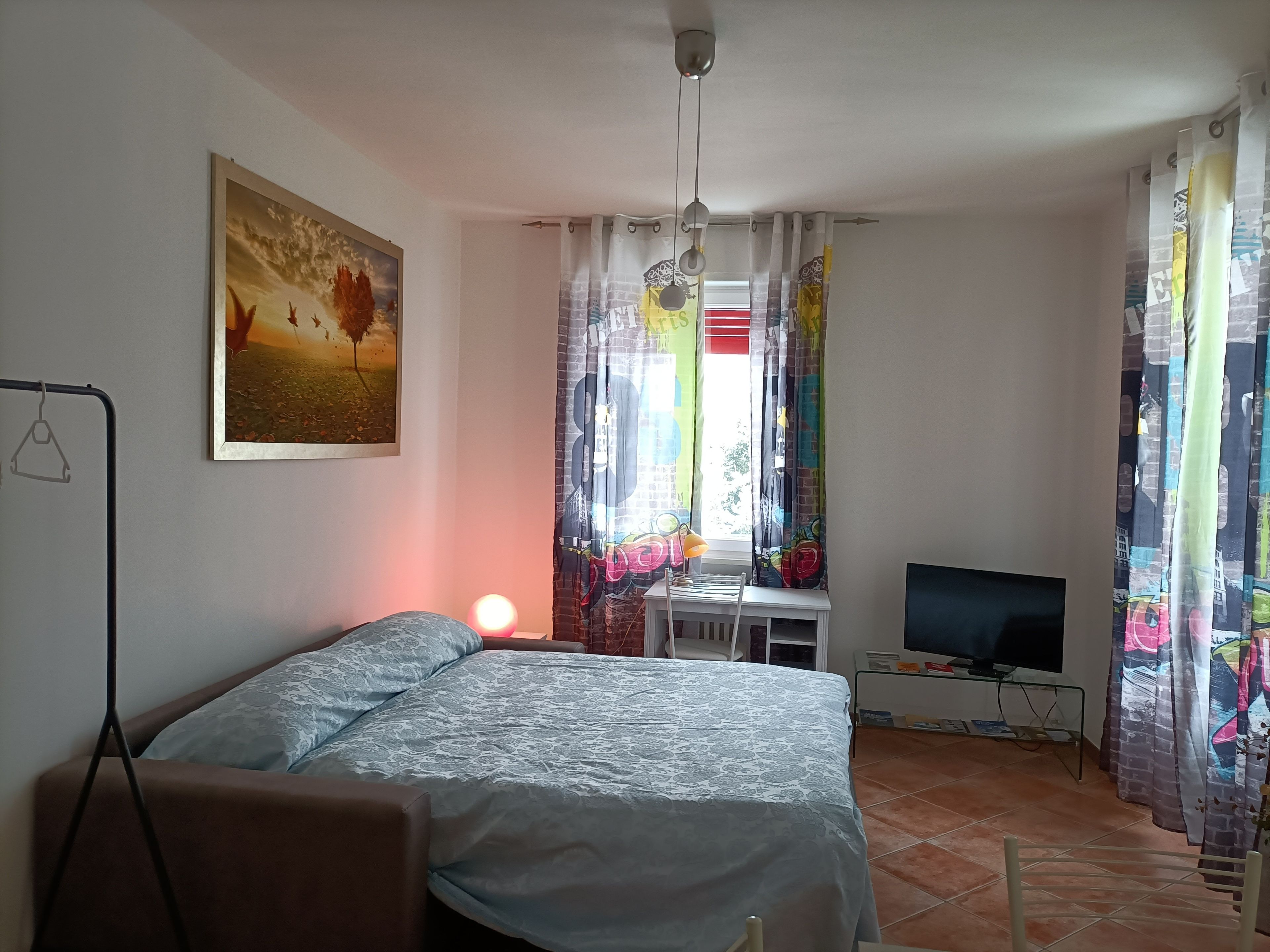 A bedroom with a bed, a desk with a TV, a painting on the wall, and curtains.