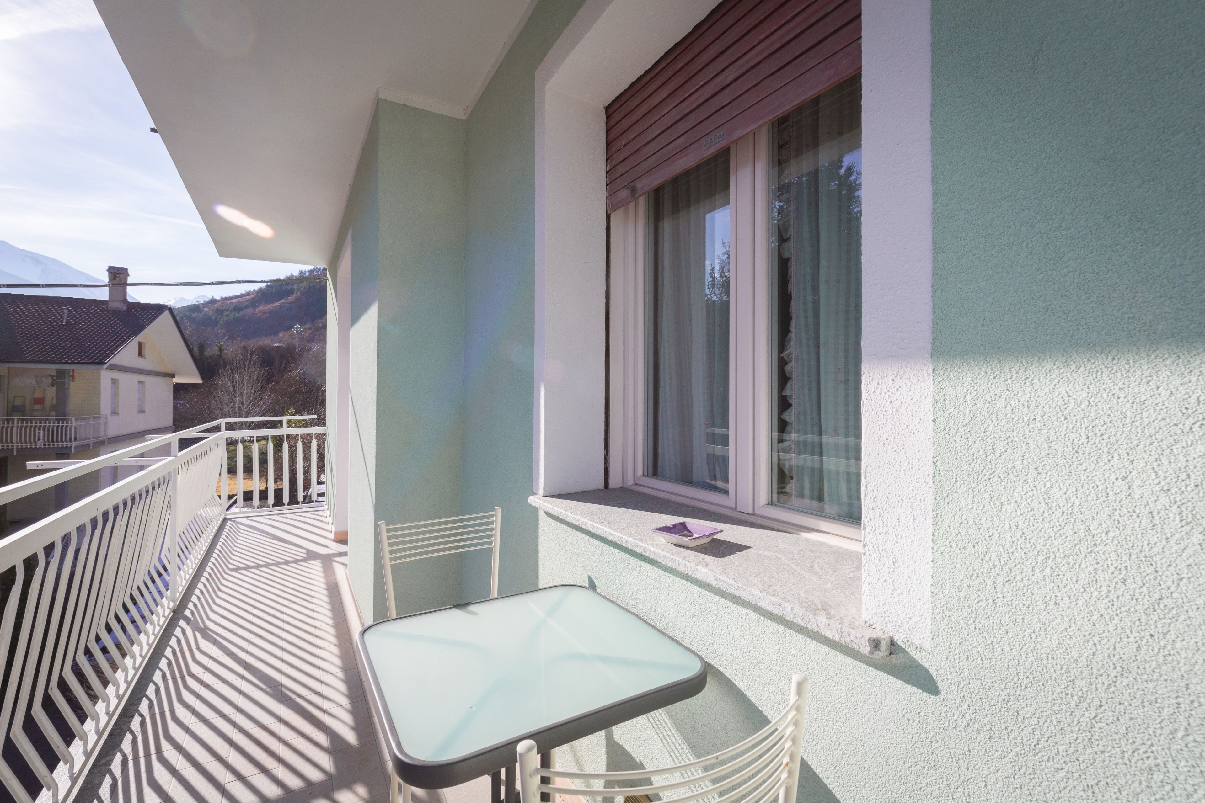 Ein Balkon mit Tisch und Stühlen, ein Fenster mit Vorhängen und Blick auf einen Berg.