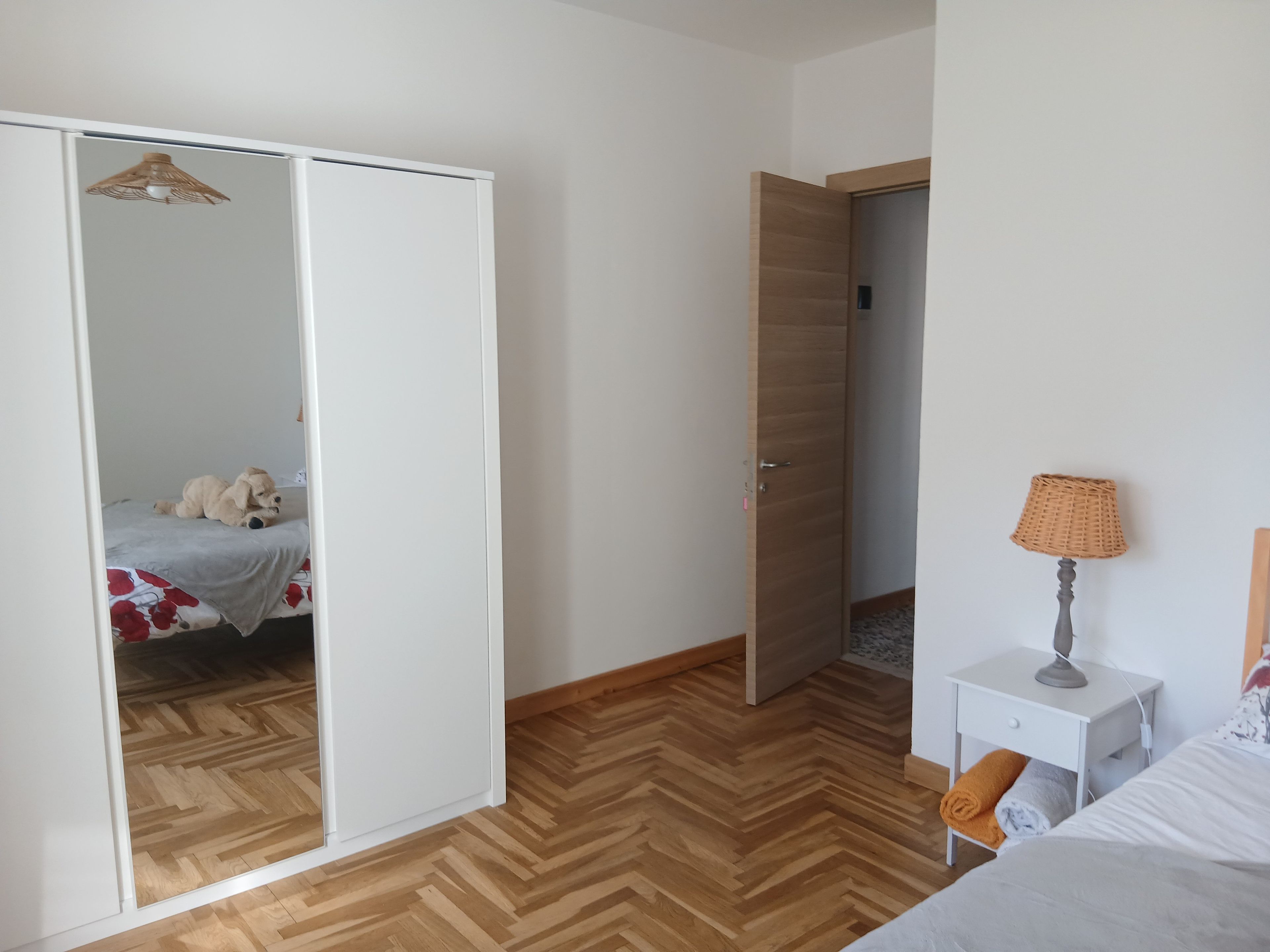 A bedroom with a wardrobe, a bed with a floral bedspread, a nightstand with a lamp, and a wooden herringbone floor.