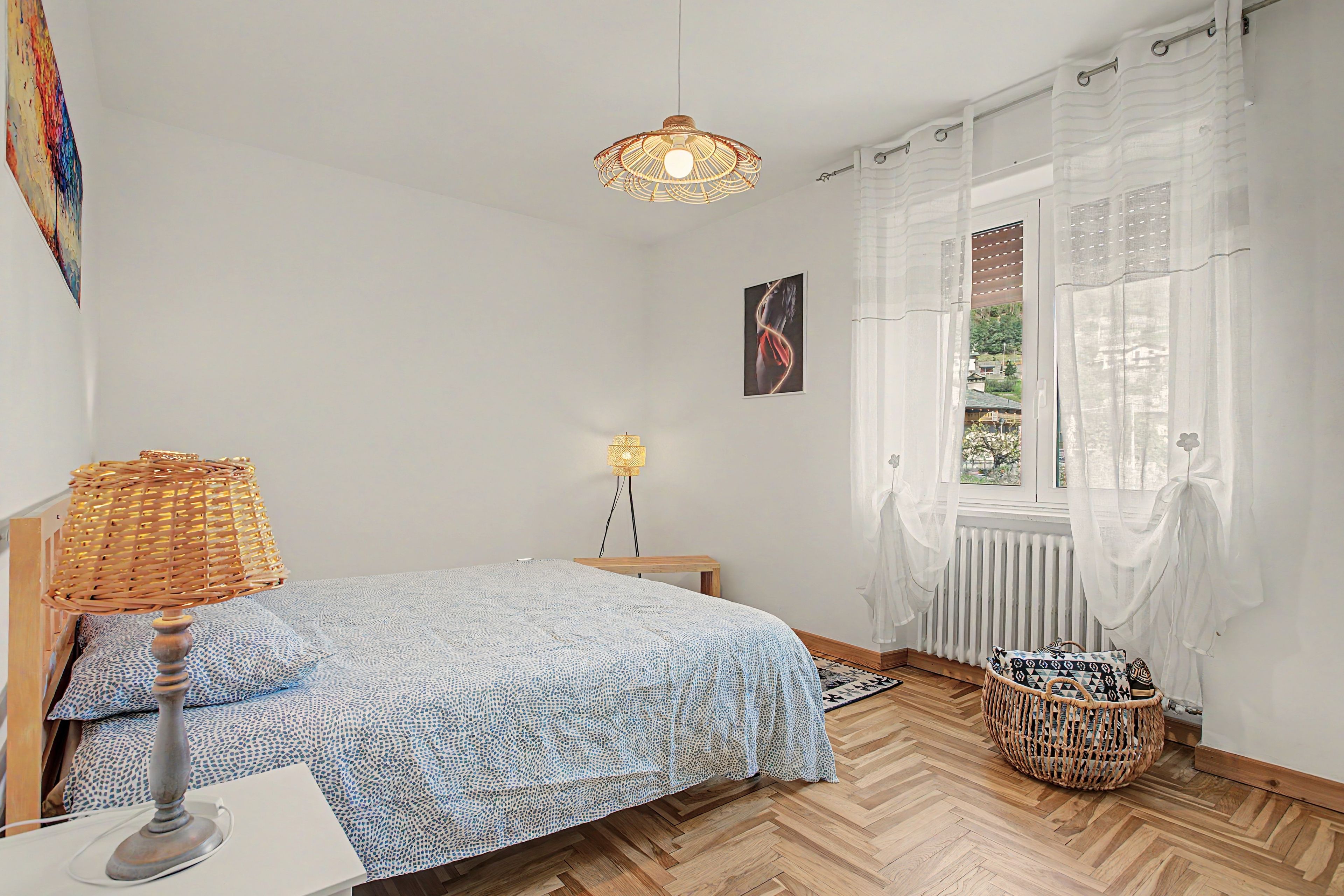 Ein Schlafzimmer mit einem Bett, einer Nachttischlampe, einer Pendelleuchte, einem Fenster mit durchscheinenden Vorhängen und einer Heizung.