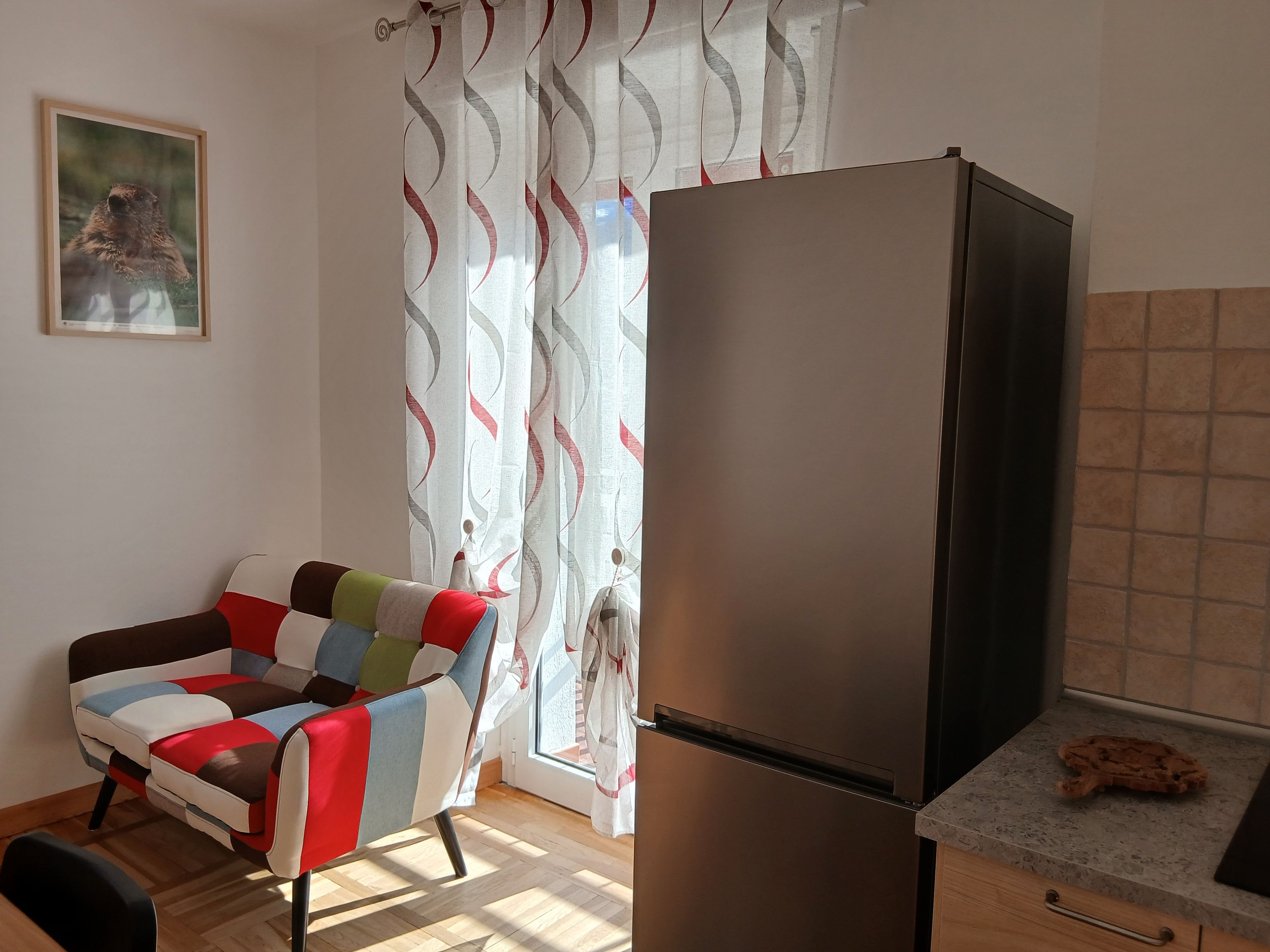 A room with a modern kitchen, a colorful armchair, a refrigerator, and a patterned curtain.