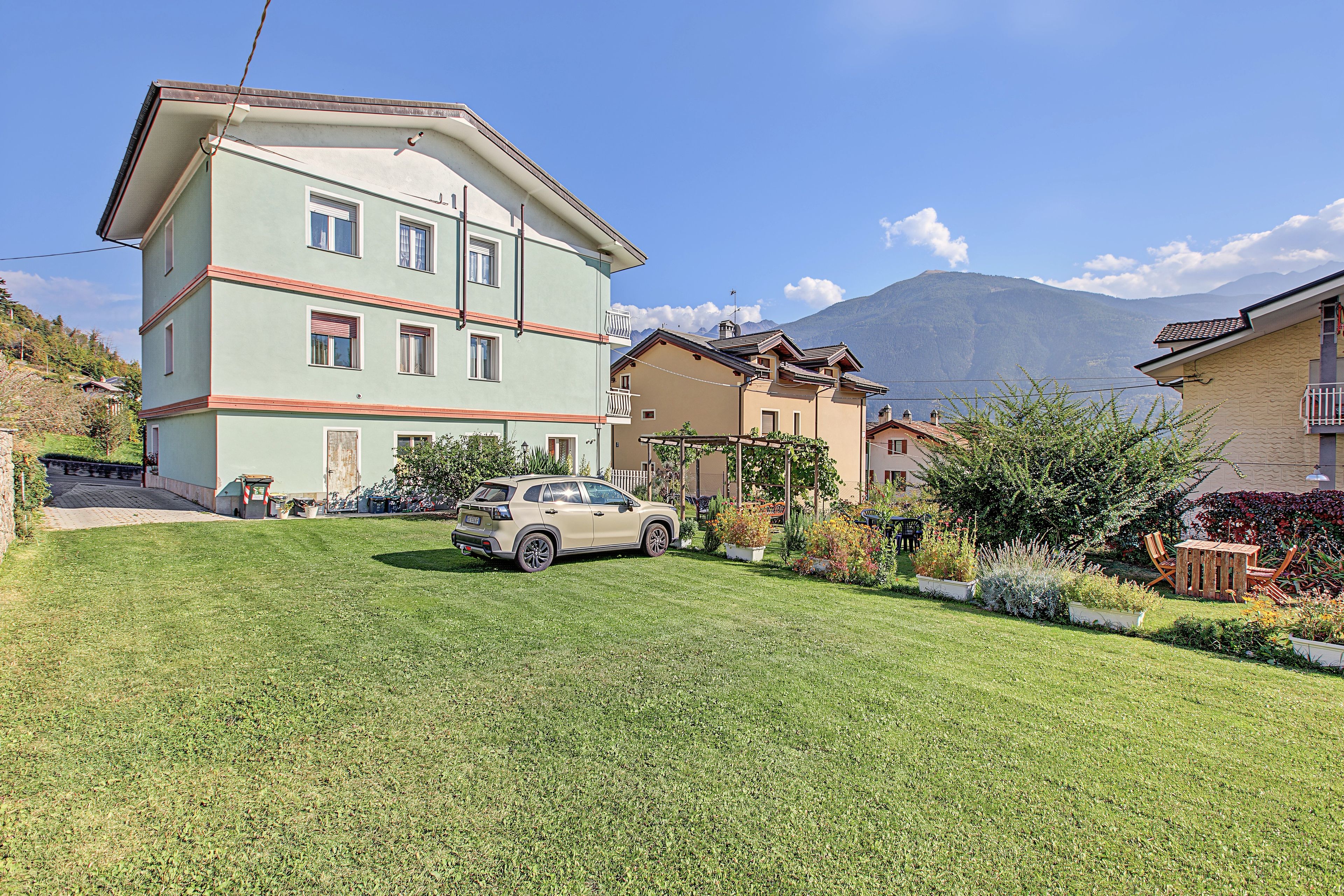 Un immeuble résidentiel à plusieurs étages, avec une voiture garée dans l’allée, entouré de jardins bien entretenus et avec une montagne en arrière-plan.