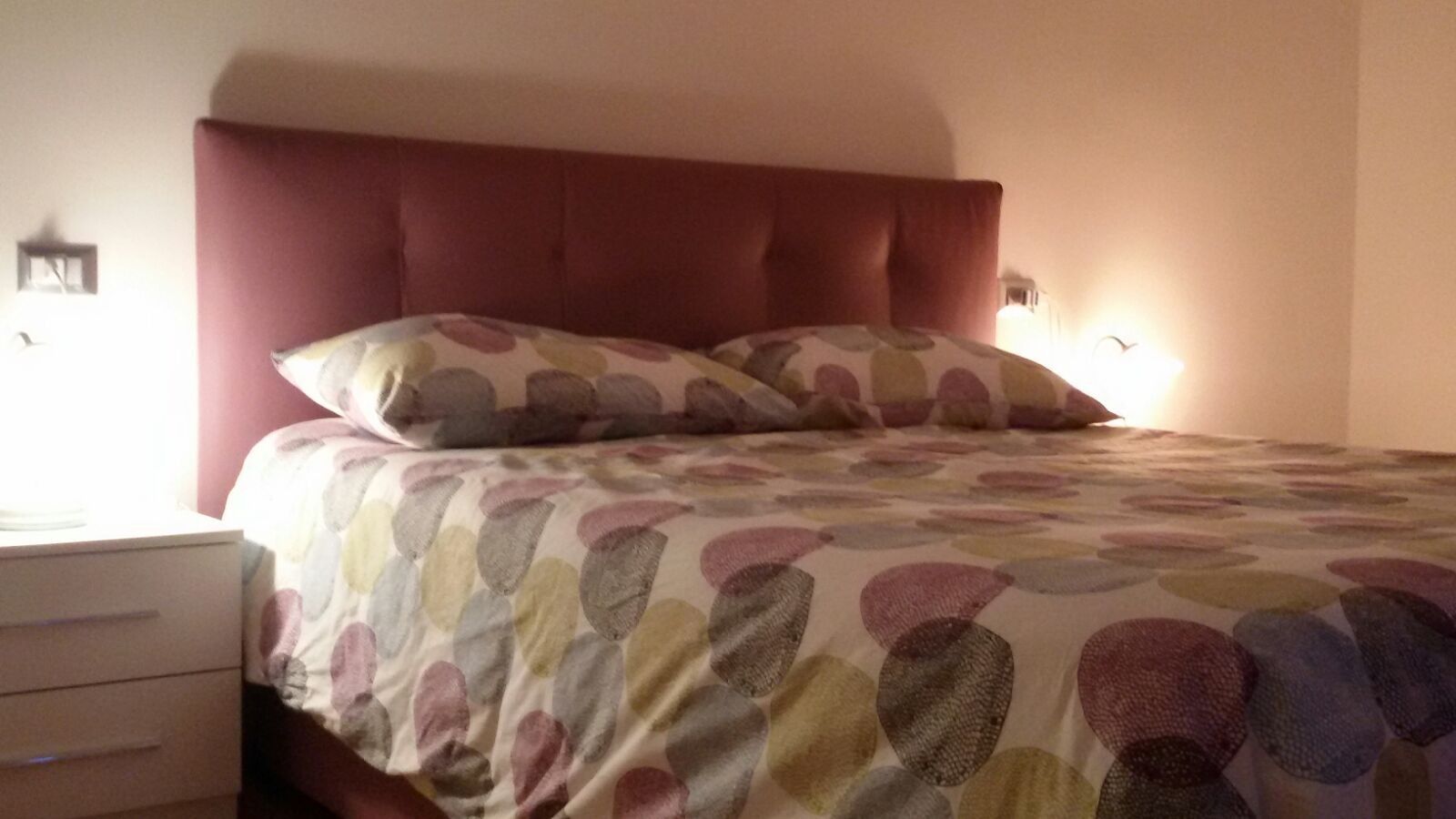A bed with a patterned bedspread and a headboard, a bedside table, and a wall-mounted lamp.