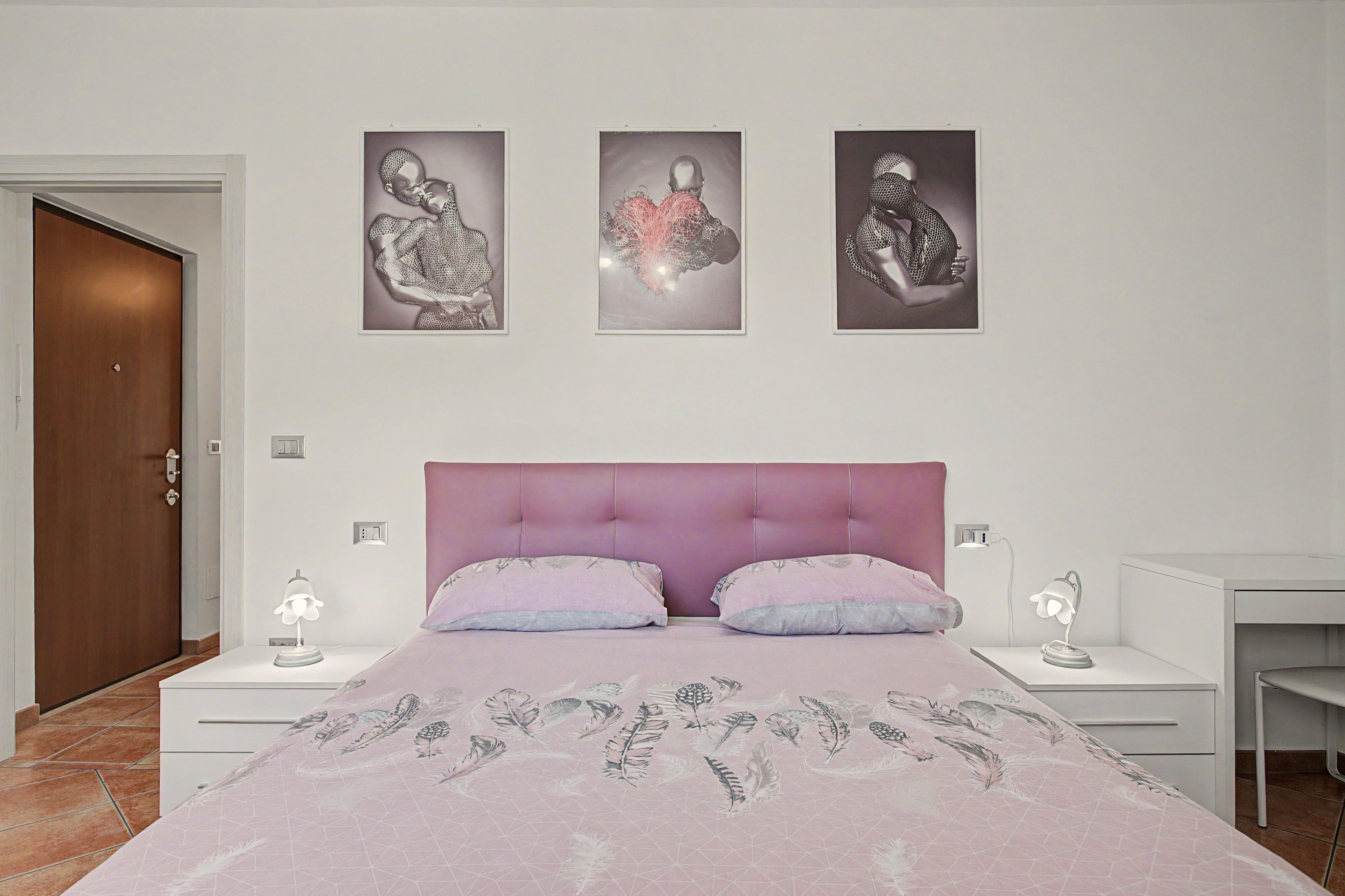Ein Schlafzimmer mit einem Bett, zwei Nachttischen, einem kleinen Tischchen, einer Tür und drei an der Wand befestigten Kunstwerken.