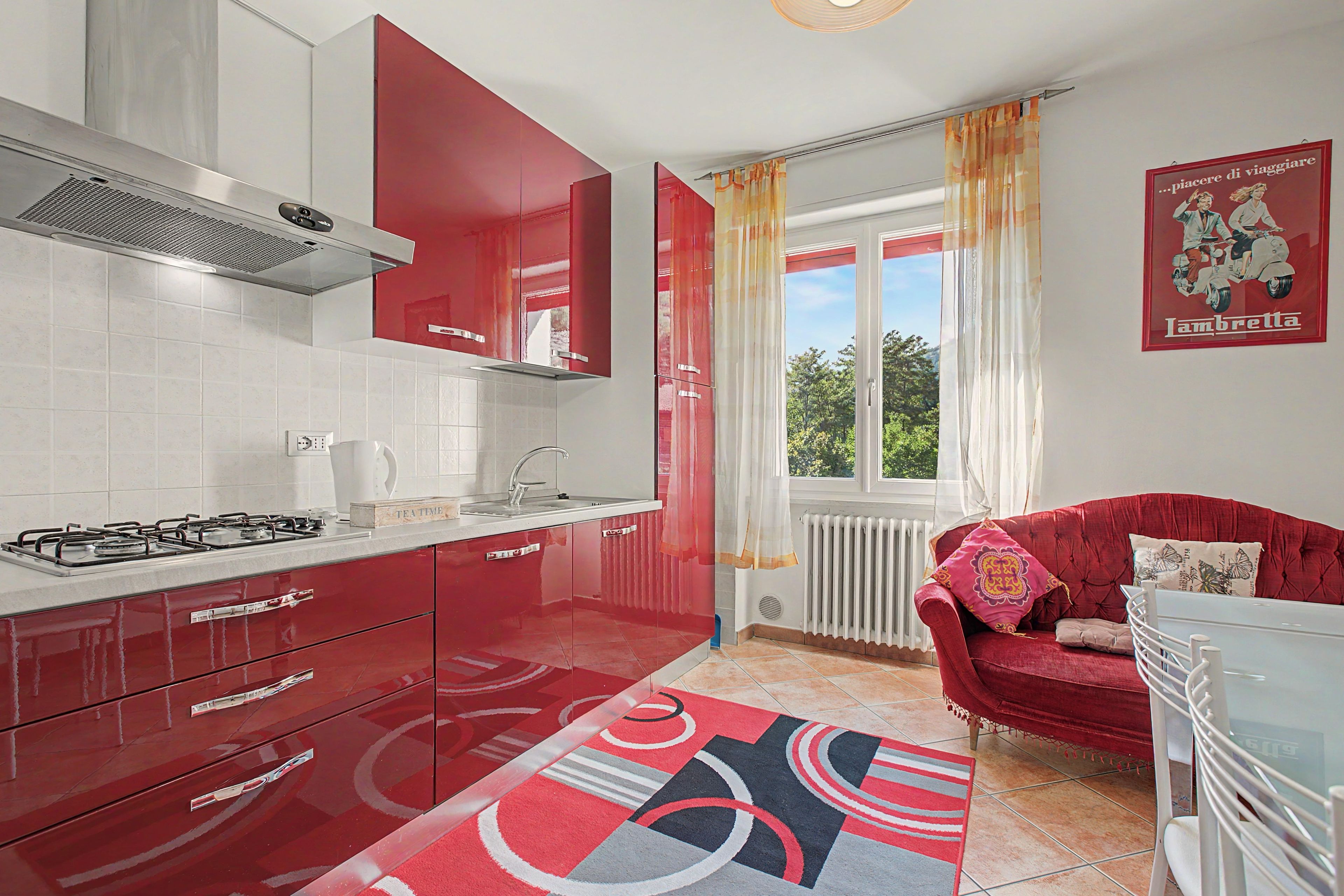 Cocina compacta con armarios rojos, estufa a gas y ventana con cortinas.