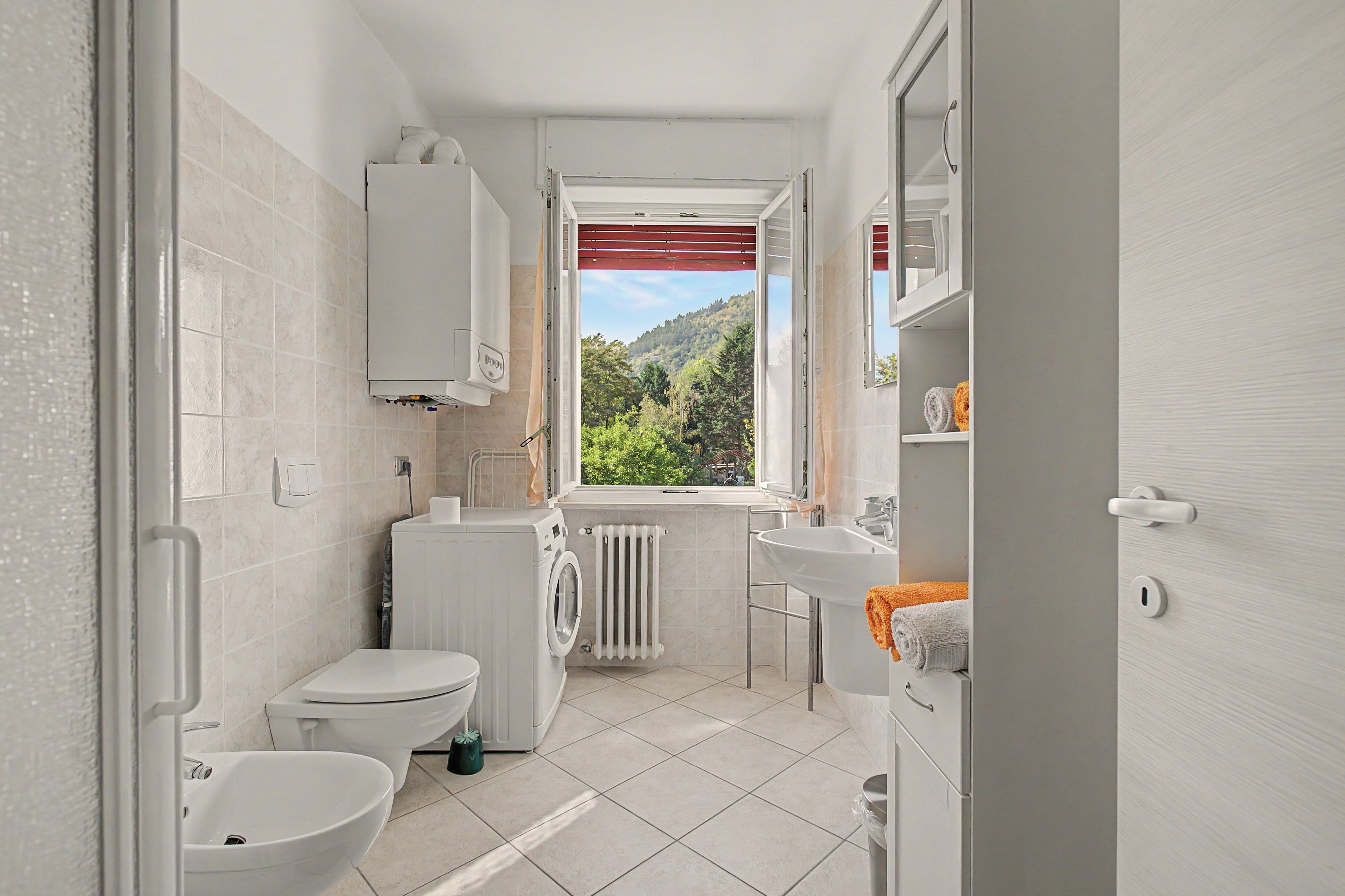 Ein kleines Badezimmer mit Toilette, Bidet und Waschbecken, einer Waschmaschine und einem Fenster mit Blick auf ein bewaldetes Gebiet.