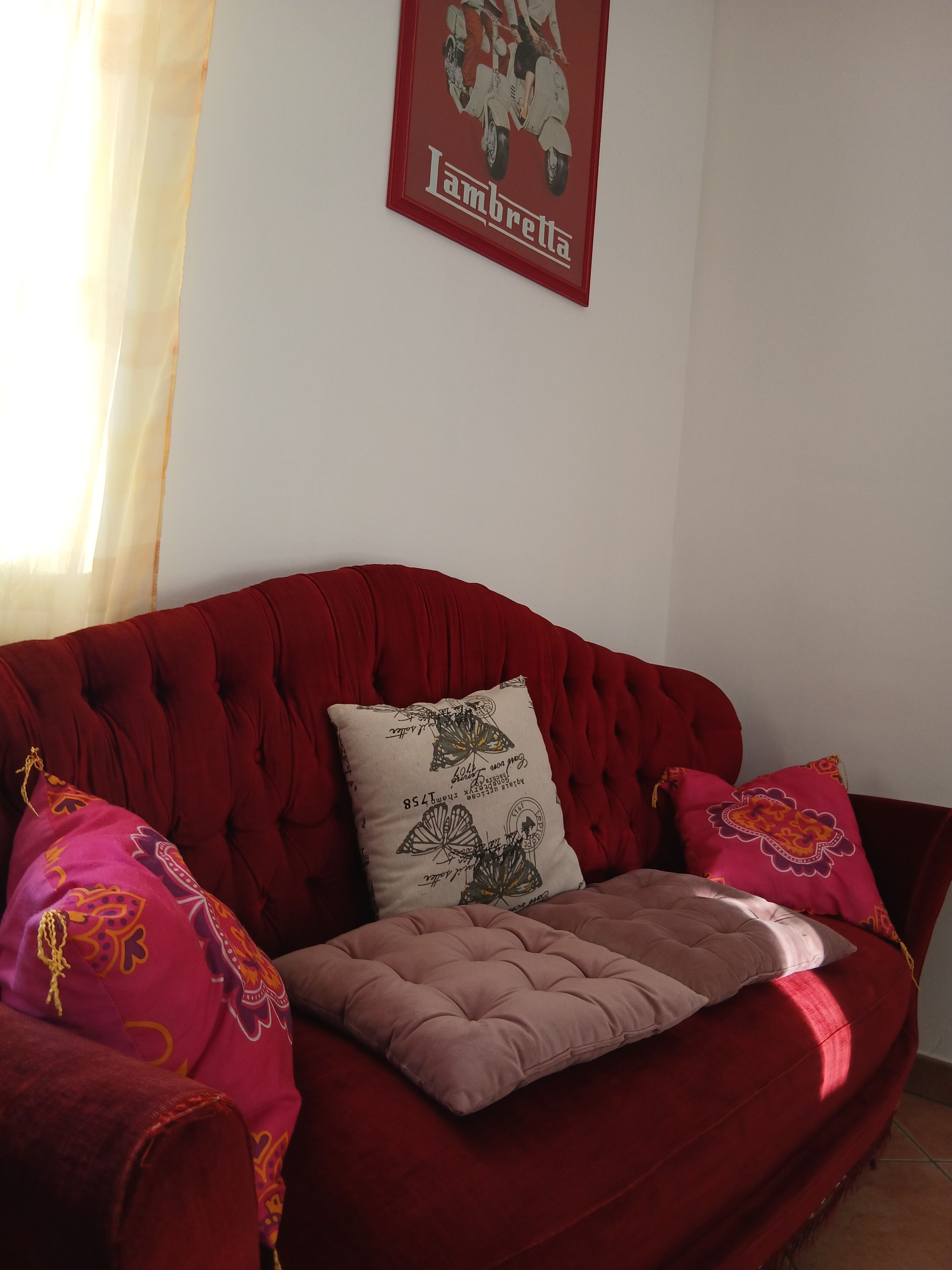 Un sofá rojo con un cojín y dos almohadones decorativos, una pared con un cuadro enmarcado y una cortina translúcida que cubre parcialmente una ventana.