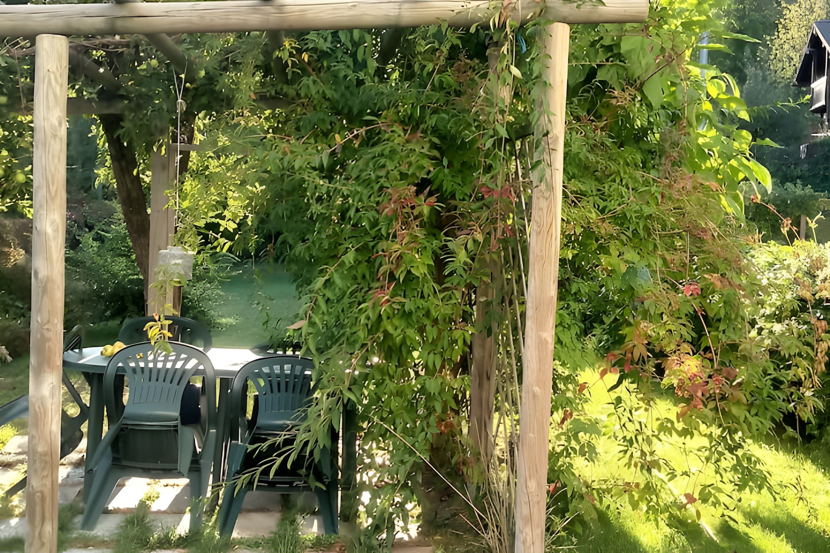 A wooden pergola with a table and chairs underneath, surrounded by lush greenery and a garden.