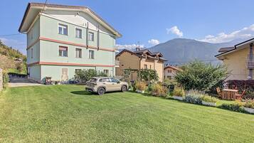 Un immeuble résidentiel à plusieurs étages, avec une voiture garée dans l’allée, entouré de jardins bien entretenus et avec une montagne en arrière-plan.