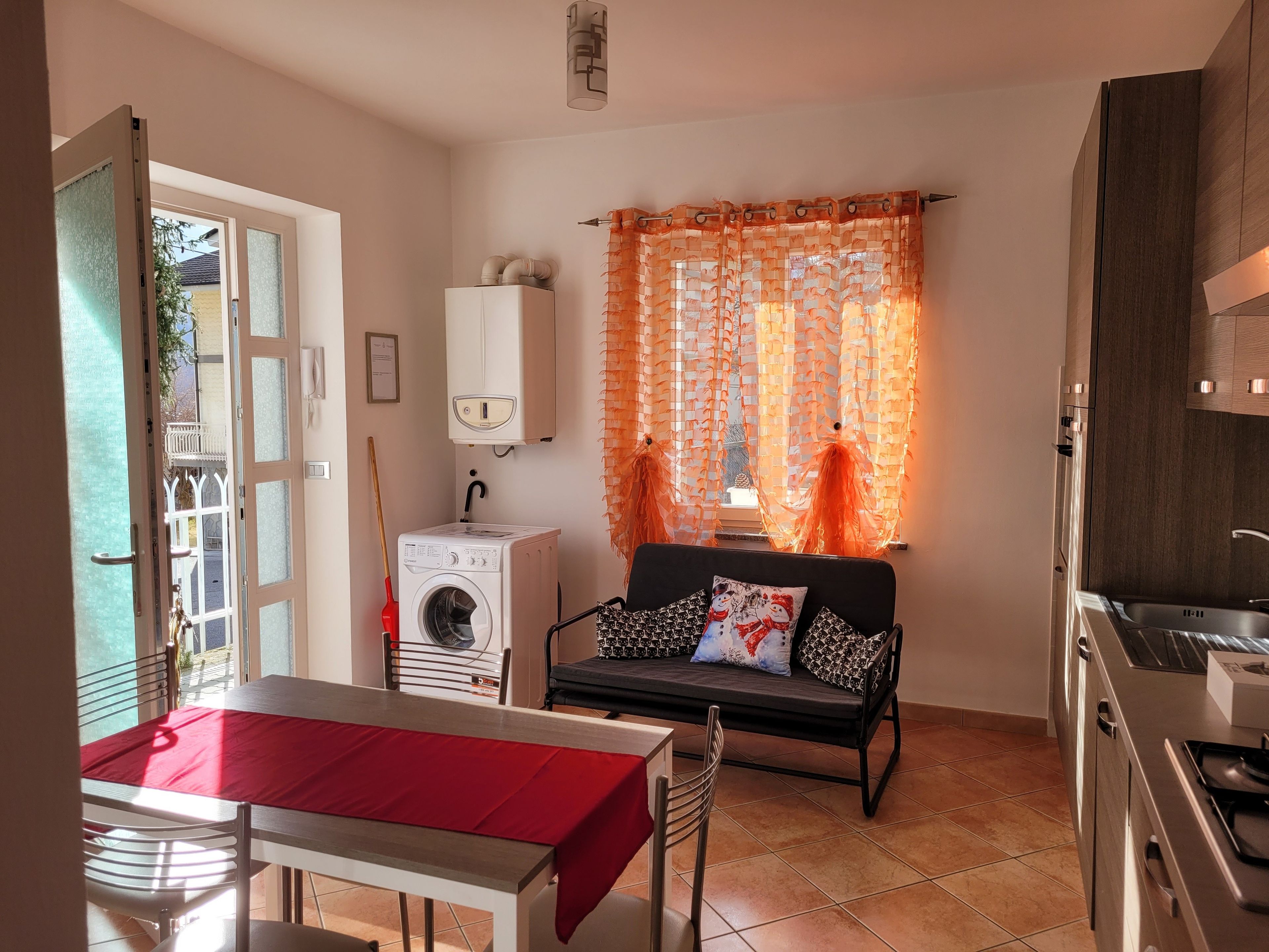A compact kitchen with a washing machine, a dining area with a red table, and a sofa with patterned cushions.