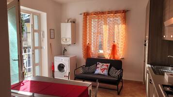 A compact kitchen with a washing machine, a dining area with a red table, and a sofa with patterned cushions.