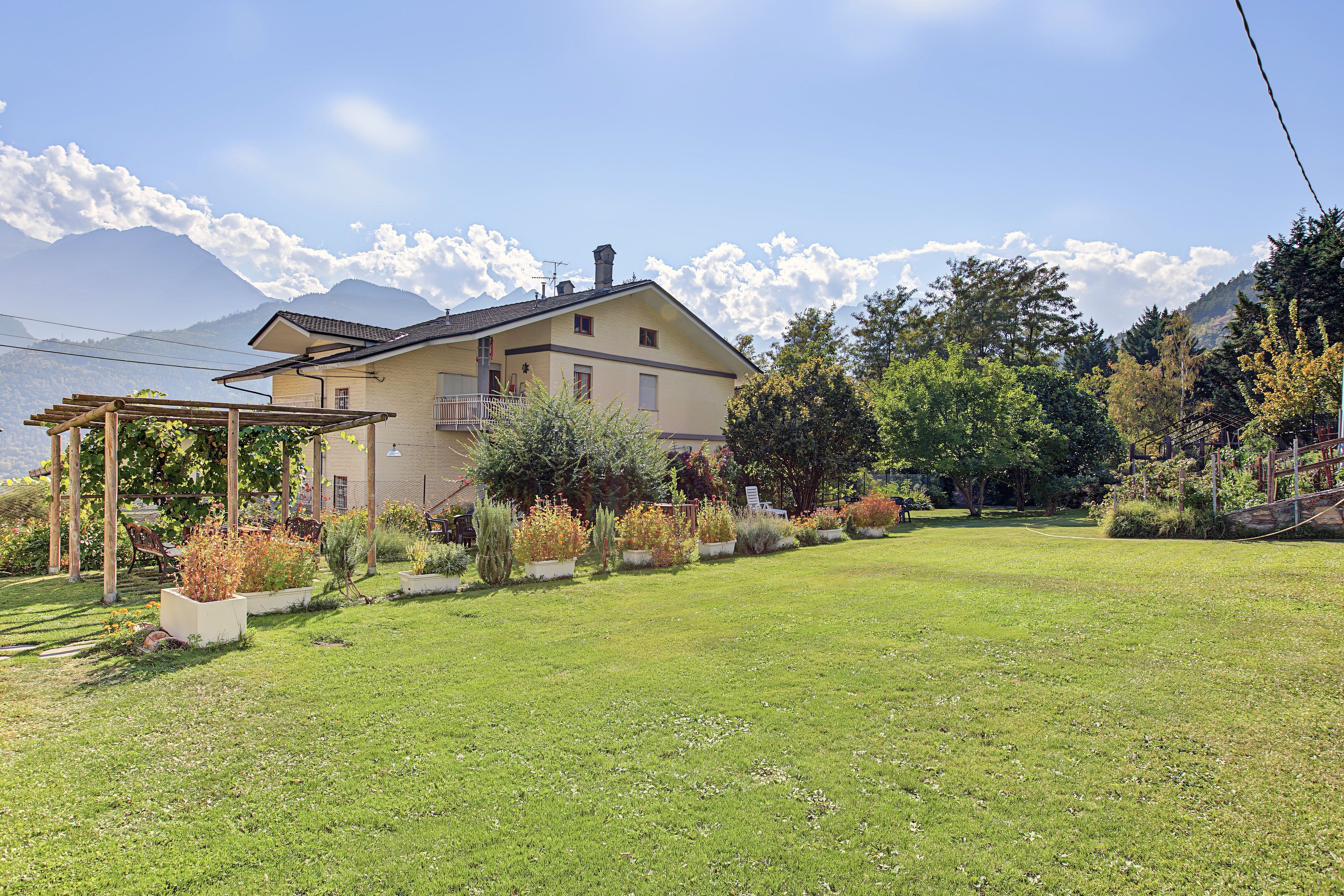 Ein Haus mit Garten, einer Pergola und Bergen im Hintergrund.