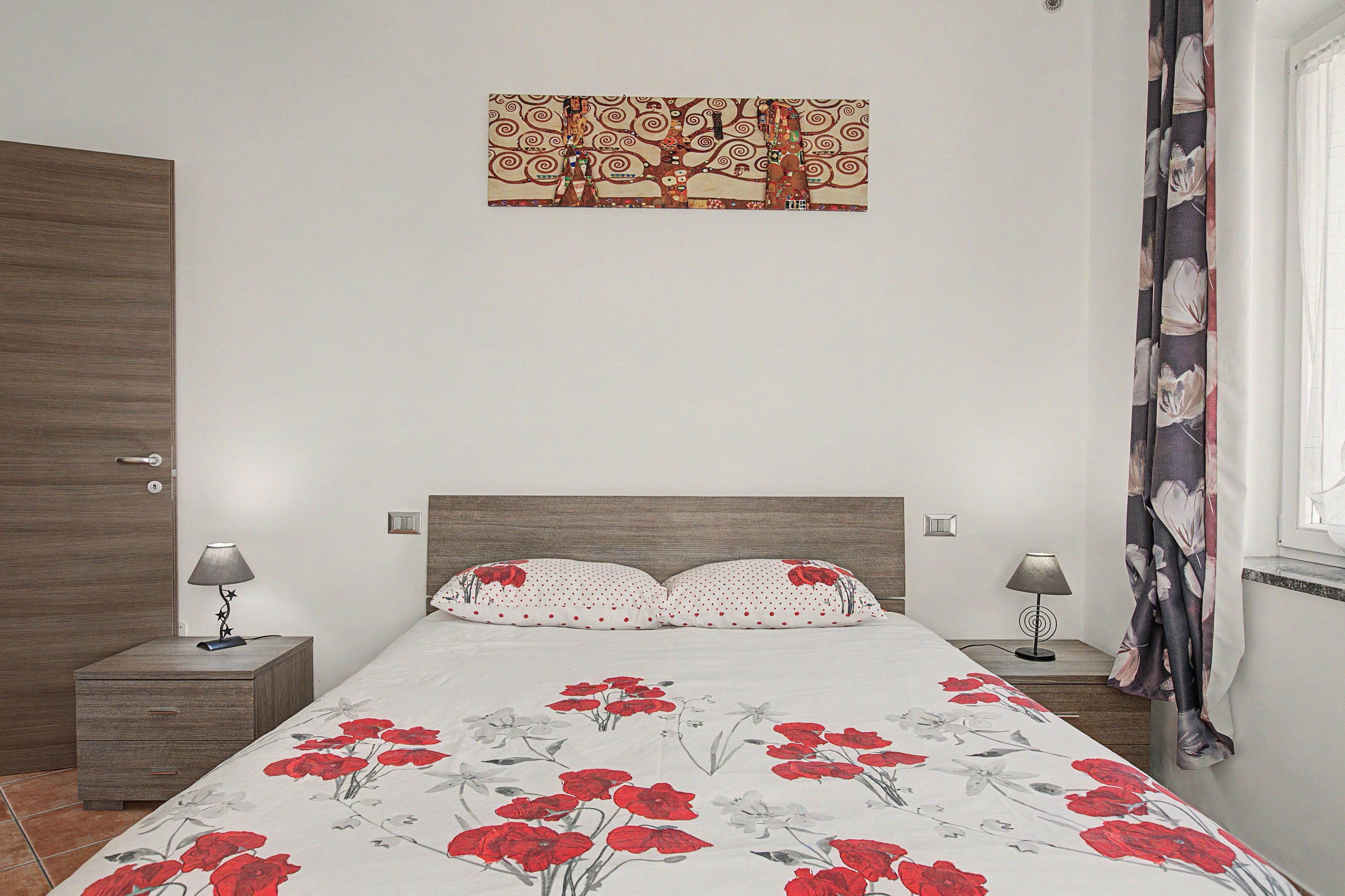 Une chambre à coucher comprenant un lit, deux tables de chevet, une commode, une œuvre d’art accrochée au mur et une fenêtre avec des rideaux.