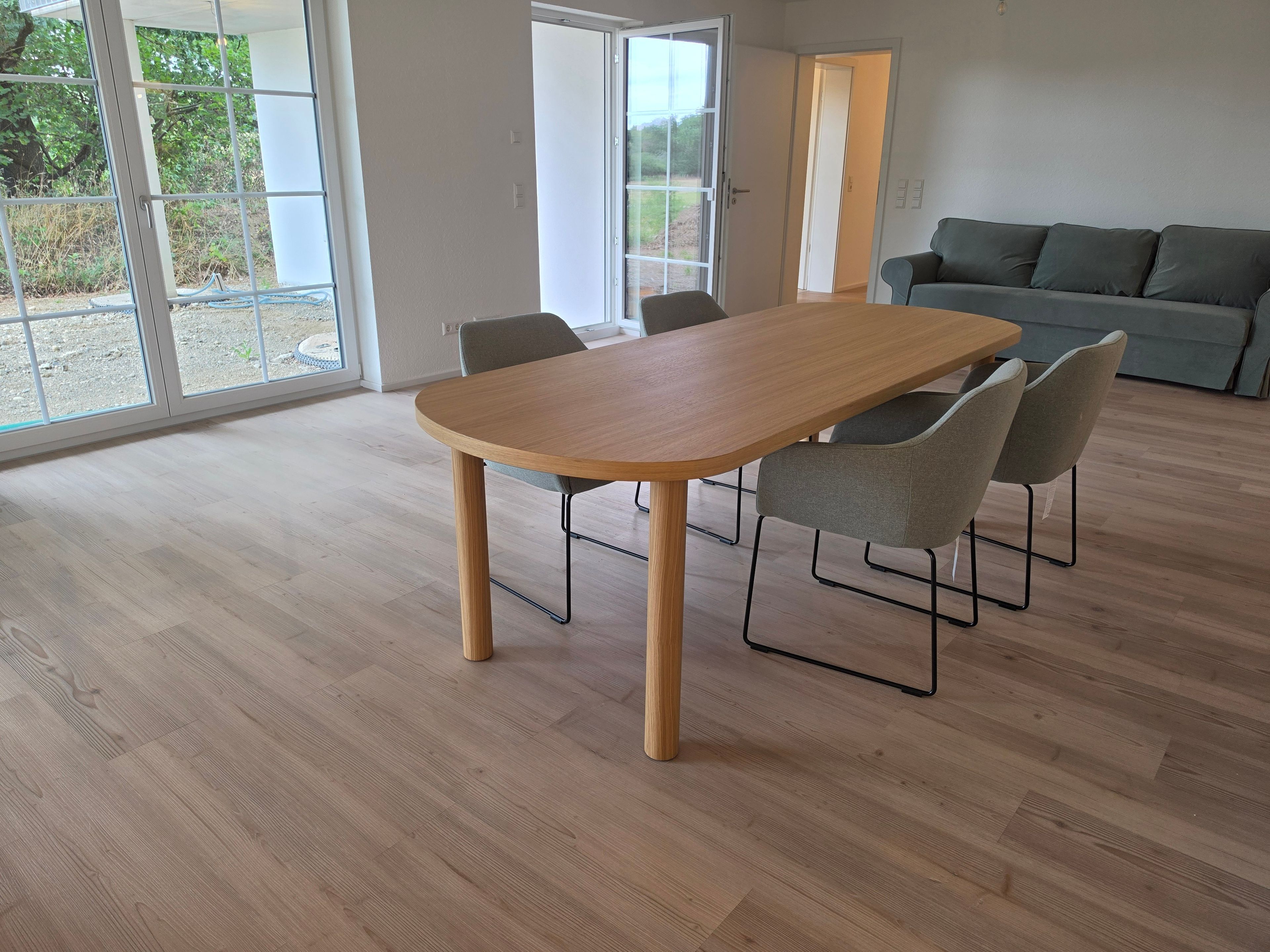 A modern living room with a wooden dining table, grey chairs, a grey sofa, and large windows.
