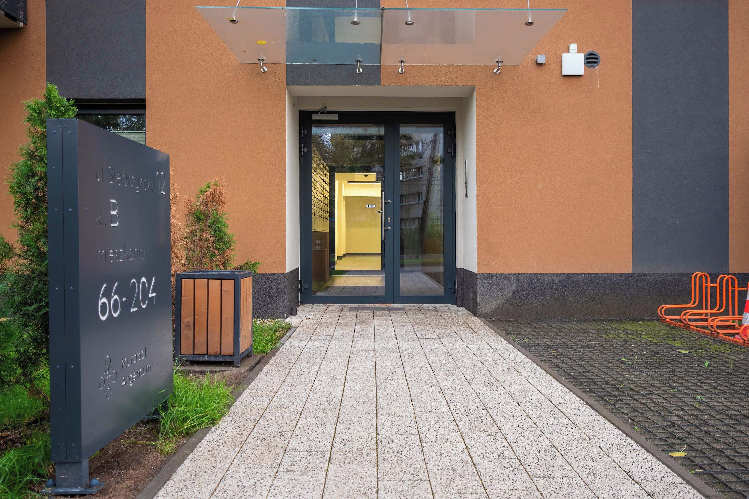 Een gebouw-ingang met een glazen luifel, een metalen bord met een nummer en een geplaveid voetpad.