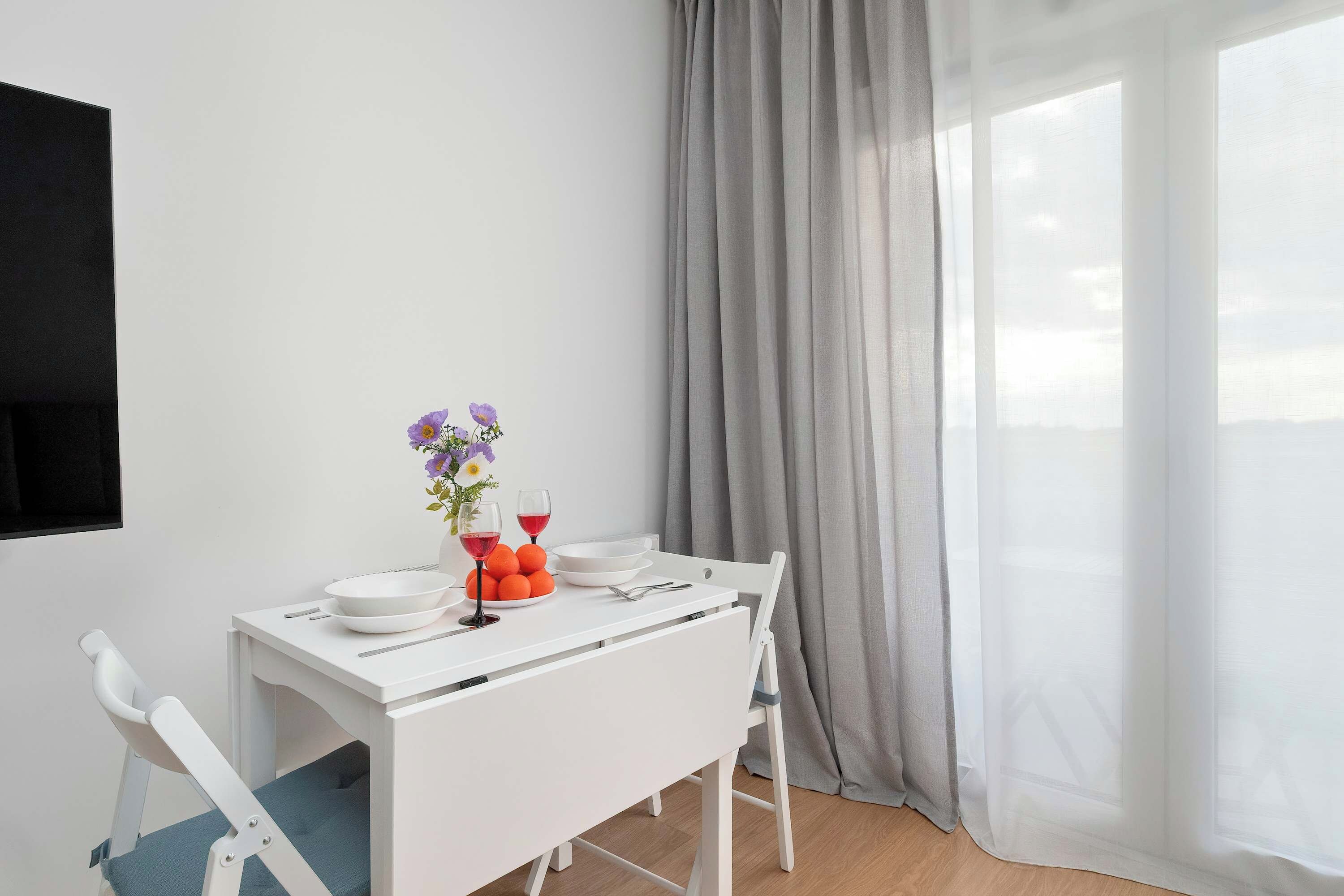 Un coin repas avec une table blanche dressée pour deux personnes, un vase rempli de fleurs et une assiette d’oranges.