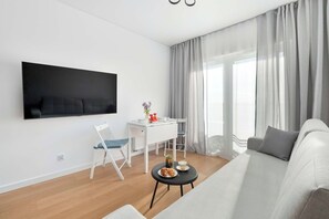 Een moderne woonkamer met een flatscreen televisie aan de muur, een wit bureau met stoel, een ronde salontafel met gebak en een grijze bank.