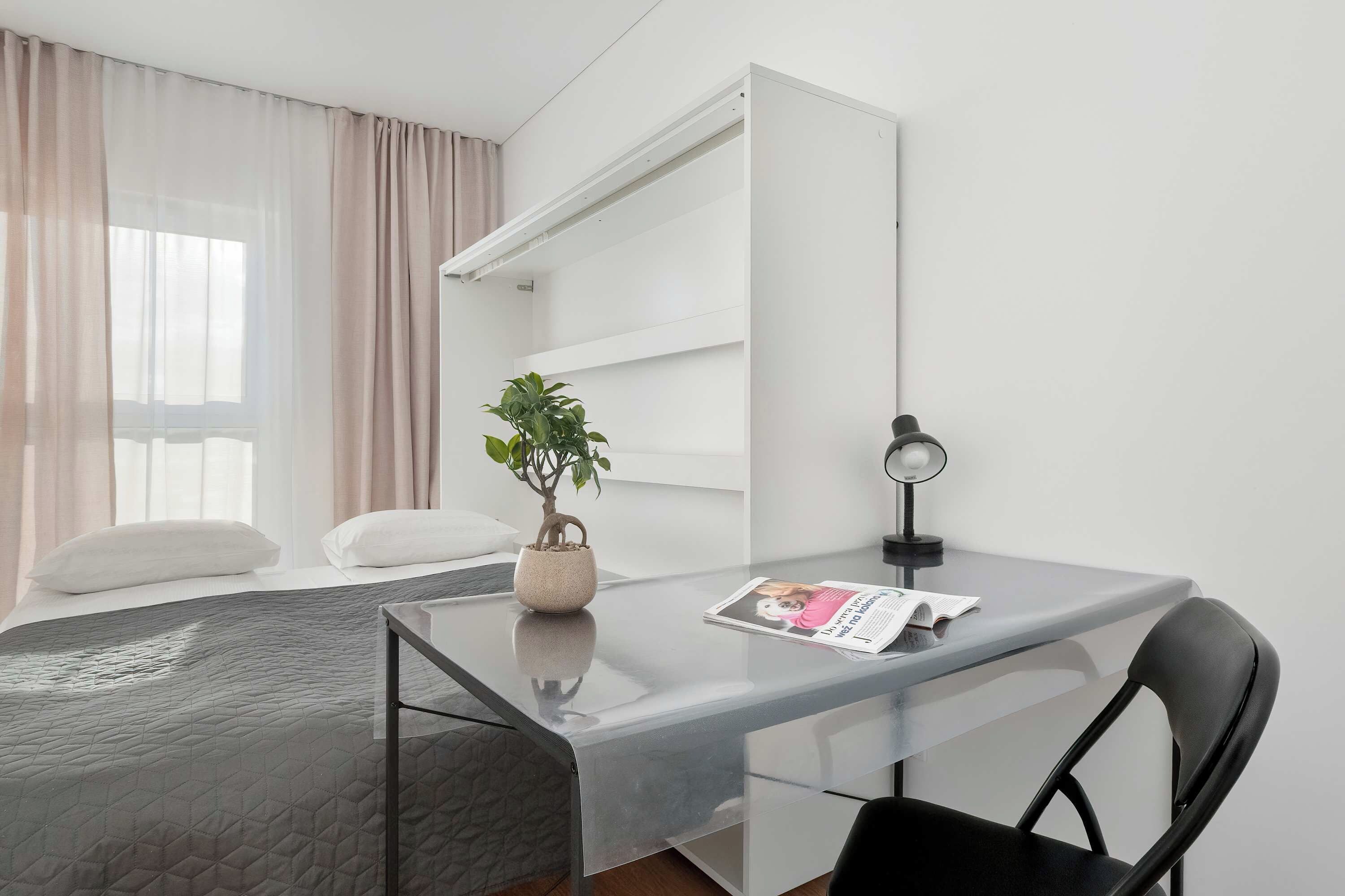 Een moderne hotelkamer met een glazen bureau, een enkele stoel, een potplant en een bed met een grijze deken.