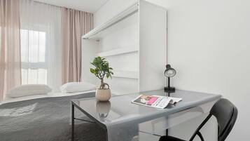Een moderne hotelkamer met een glazen bureau, een enkele stoel, een potplant en een bed met een grijze deken.